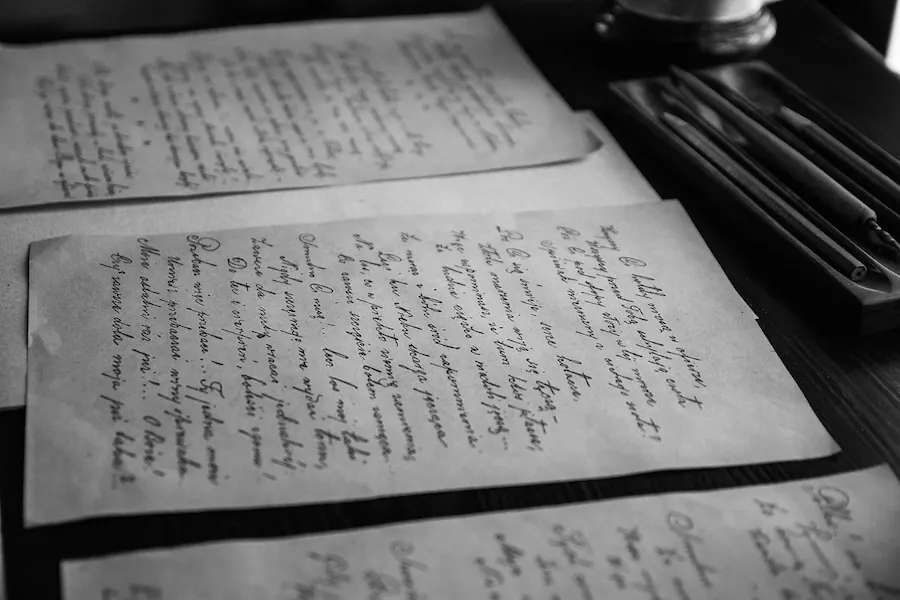 Several sheets of handwritten letters laid across a dark wooden table beside a tray of pens